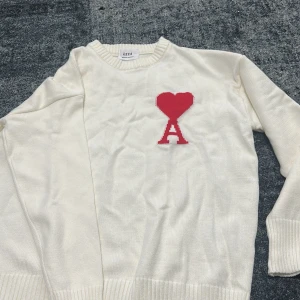 Vit stickad tröja från Ami Paris - Vit stickad tröja från Ami Paris med röd hjärtlogga och bokstaven A på bröstet. Tröjan har rund halsringning, ribbade muddar och lång ärm. Perfekt för dig som gillar stilrena plagg med en twist.