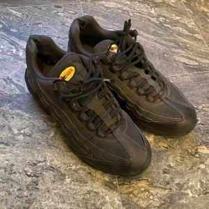 Nike Air Max 95 svarta sneakers - Nike Air Max 95 sneakers i svart med gula detaljer på tungan och sulan. Skorna har en chunky siluett, synliga Air-enheter i sulan och snörning framtill. Ovandelen är i mesh och syntet med vågiga paneler och rund tå. Perfekt för dig som gillar streetwear och sportig stil.