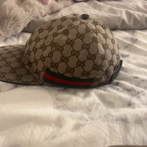Gucci keps med GG-mönster - Snygg keps från Gucci i beige och brunt med klassiskt GG-monogram över hela kepsen. Har justerbart spänne bak i svart skinn och en ikonisk grön-röd rand på sidan. Perfekt accessoar för att lyfta din outfit.