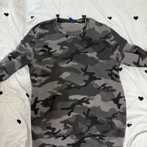 Camo-mönstrad sweatshirt i grå toner - Säljer en snygg sweatshirt med camouflage-mönster i olika grå och svarta nyanser. Tröjan har rund halsringning och långa ärmar, perfekt för dig som gillar streetstyle och vill sticka ut lite extra.