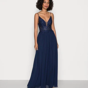 Marinblå långklänning med spets - Säljer en marinblå långklänning med smala axelband och djup v-ringning. Överdelen har vacker spets och paljettbroderi, medan kjolen är i ett lätt, flödande material. Klänningen har öppen rygg och markerad midja, perfekt för bal eller student.