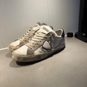 Philippe Model vita sneakers med svart detalj - Säljer ett par snygga Philippe Model sneakers i vitt med grå mockadetaljer och svart häl. Klassisk låg modell med rund tå, snörning och platt sula. Skorna har en ikonisk sköld på sidan och är tillverkade i skinn och mocka för en cool look.