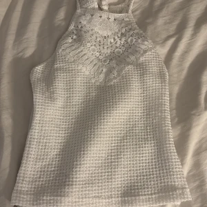 Vit topp med spets - Säljer en vit linnetopp med snygg spetsdetalj upptill och strukturerad yta. Toppen är ärmlös och har en halterneck-design som knyts💕