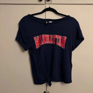 Mörkblå Manhattan knyt-topp - Säljer en mörkblå topp med röd och vit Manhattan-tryck framtill. Toppen har korta ärmar, rund hals och en snygg knytning längst ner som ger en chill och trendig vibe. Perfekt för dig som gillar streetstyle och college-inspirerat mode.