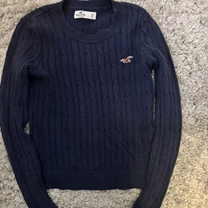 Mörkblå stickad tröja Hollister XS - Mörkblå stickad tröja från Hollister med klassiskt kabelstickat mönster och rund hals. Tröjan har lång ärm och en broderad Hollister-logga på bröstet. Perfekt för lager-på-lager och chill höst- och vinterstil.