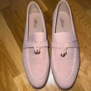 Rosa mockaloafers från Mulanka - Snygga rosa loafers från Mulanka i storlek 41. Skorna är gjorda i mjuk mocka med dekorativ metallring framtill och har en klassisk rund tå. Perfekt för dig som gillar stilrena och trendiga detaljer i din outfit.