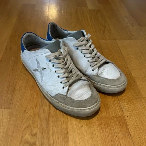 Axel Arigato vita sneakers med blå detalj - Snygga vita sneakers från Axel Arigato med blå häl och grå mockadetaljer. Levereras med originalkartong och dustbag.
