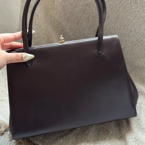 Vintage Mörkbrun handväska i skinn med gulddetaljer - Snygg mörkbrun handväska i slätt skinn med stilrena handtag och guldfärgade metalldetaljer på sidorna och låset. Väskan har en klassisk, boxig form och passar perfekt för dig som gillar retrovibbar och elegant design.