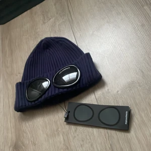 Mörkblå C.P. Company Goggle mössa / helt ny  - Säljer en mörkblå ribbstickad Goggle mössa från C.P. Company med ikoniska svarta glasögon-detaljer framtill. Mössan är tillverkad i mjukt stickat material och har en bred uppvikt kant. Perfekt för dig som vill sticka ut med streetwear-vibe.