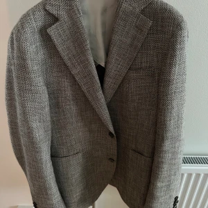 Oscar Jacobson kavaj - Snygg grå kavaj från Oscar Jacobson, tillverkad i Italien. Modellen heter Ferry Patch Soft Blazer och har en klassisk passform med två knappar framtill, fickor och diskret struktur i tyget. Perfekt för dig som gillar stilrena och tidlösa plagg. 