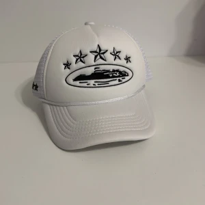Vit Corteiz truckerkeps med stjärnor - Snygg vit truckerkeps från Corteiz med svart broderade stjärnor och logga framtill. Kepsen har mesh på sidorna och bak, samt justerbar snapback. Märkesband på insidan och broderade detaljer på sidan. Perfekt för dig som vill sticka ut med streetwear.