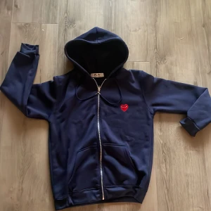 Mörkblå hoodie Comme des Garçons Play - Säljer en mörkblå hoodie från Comme des Garçons Play med dragkedja och huva. Tröjan har det ikoniska röda hjärtat broderat på bröstet och praktiska fickor framtill. Perfekt för dig som gillar streetstyle och vill ha något stilrent men ändå lekfullt.