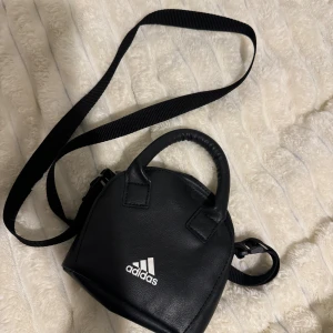 Svart axelväska från Adidas - Snygg svart axelväska från Adidas med vit logga framtill. Väskan har både handtag och justerbar axelrem, perfekt för att bära med sig det viktigaste. Tillverkad i skinnimitation med stilren design och kompakta mått.