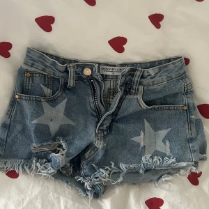 Blå lågmidjade  jeansshorts med stjärnor💖 - Säljer ett par blå jeansshorts från Design By Amor med ljusa stjärnor som mönster. Shortsen har fransig och sliten kant, klassisk femficksdesign och dragkedja med knapp. Perfekta för dig som gillar en cool och avslappnad stil. 