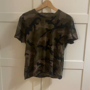 Camouflage t-shirt från Valentino - Snygg camouflage t-shirt från Valentino, tillverkad i Italien. T-shirten har klassisk rund hals och korta ärmar. Färgerna går i grönt, brunt och svart med ett coolt camomönster. Materialet är mjuk bomull och bak på halsen finns en liten metallnit som detalj.