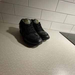 Valentino Garavani svarta sneakers - Svarta sneakers från Valentino rockrunners med coolt camouflage-mönster och snörning. Skorna har en rund tå och platt sula, samt detaljer i både mocka och skinn. Tungan har en olivgrön logotyp. Perfekt för dig som gillar exklusiva streetwear-vibes.