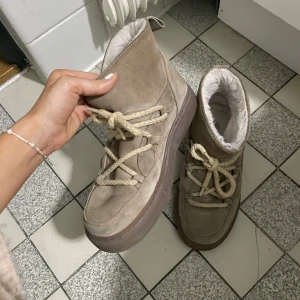 Beige Inuikii boots med snörning - Säljer ett par beige boots från Inuikii med grov snörning och platt brun sula. Utsidan är i mjuk mocka och insidan är fodrad med fluffigt material för extra värme. Skorna har rund tå och en snygg detalj med logotypen broderad bak. Obs!! endast använda en halv vinter och jag har tagit av metallgrejen 