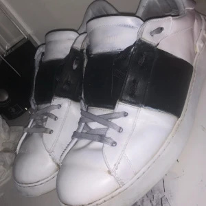 Valentino Open sneakers vit/svart - Säljer ett par Valentino Open sneakers i vitt skinn med bred svart läderdetalj över mitten. Skorna har rund tå, platt sula och grå snörning. Snygg och stilren design som sticker ut med den kontrasterande svarta panelen. Perfekta för dig som gillar exklusiva sneakers.