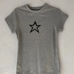 Grå t-shirt med svart stjärna - Snygg grå t-shirt med svart stjärntryck på bröstet. Modellen har rund hals och korta ärmar, perfekt för en chill och enkel stil. Materialet känns mjukt och skönt mot huden, och passformen är klassisk utan att vara för tight. Gjorde den på syslöjden
