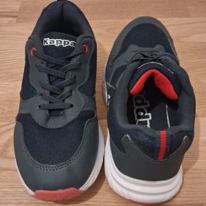 Svarta sneakers från Kappa strl 37 - Säljer ett par svarta Kappa sneakers med vita och röda detaljer. Skorna har snörning, rund tå och platt sula. Ovandelen är i syntet och textil, med Kappa-logga på sidan. Insidan har röd vaddering för extra komfort. Perfekt för dig som gillar sportig stil.
