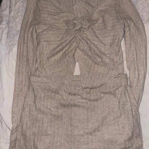 Beige ribbad kort kjol o topp med knyt - Trendig beige ribbad kort kjol  med långärmad topp och snygg knytning framtill. Toppen har v-ringning, markerad midja och en liten slits framtill. Materialet är mjukt och stretchigt, perfekt för en cool och bekväm look.