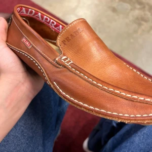 Bruna loafers i skinn från Prada - Snygga bruna loafers från Prada i mjukt skinn med kontrasterande vita sömmar och diskret logga på sidan. Skorna har klassisk form och en bekväm passform, perfekt för dig som gillar stilrena och exklusiva detaljer. INTRESSEKOLL KÖP EJ STRL 40 MEN PASSAR 42