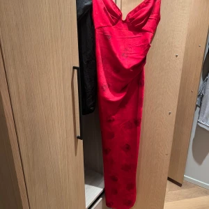 Röd långklänning med blommönster - Snygg röd långklänning i glansigt material med diskreta blommönster. Klänningen har v-ringad urringning, rynkade axelband och figurnära passform som ger en elegant look. Klänningen är använd under endast en kväll!!