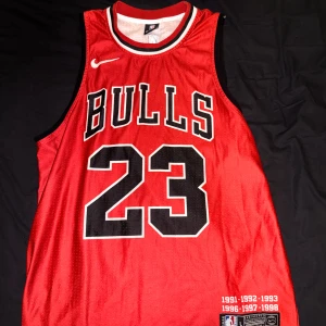 Chicago Bulls Jordan #23 baskettröja Nike - Säljer en ikonisk Chicago Bulls baskettröja med Michael Jordans nummer 23. Tröjan är röd med svarta och vita detaljer, stora tryck fram och bak, samt broderade NBA- och Nike-loggor. Tillverkad i ventilerande meshmaterial, perfekt för basket eller streetwear.