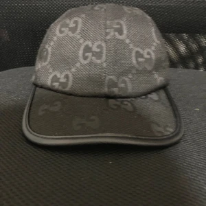 Svart keps - Snygg svart keps från med klassiskt GG-mönster över hela kepsen. Justerbar rem  Kepsen är tillverkad i slitstark polyester och har en böjd skärm. Perfekt för dig som vill ha en lyxig streetstyle-accessoar.