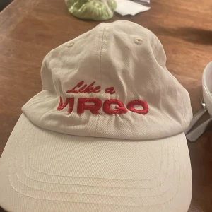 Vit keps Sarah Klang Like a Virgo - Vit keps med röd broderad text 'Like a VIRGO' framtill och 'Sarah Klang' baktill. Justerbar rem med metallspänne. Klassisk form med böjd skärm och ventilerande hål. Perfekt för dig som gillar astrologi och svensk pop.