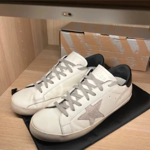 Vit sneaker från Golden Goose - Golden Goose sneakers i vitt skinn med grå mockastjärna på sidan och grå snörning. Skon har svart hälparti och rund tå, samt platt sula i gummi. Klassisk och clean design med perforerade detaljer på plösen. Perfekt för dig som gillar streetstyle.