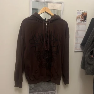 Brun hoodie med tryck och dragkedja - Säljer en brun hoodie med svart tryck framtill och dragkedja. Tröjan har huva med snörning och långa ärmar. Perfekt för dig som gillar en avslappnad och cool stil. Finns två jätte små vita fläckar som ska gå och få fort.