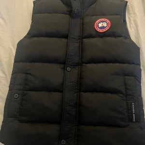 Svart dunväst från Canada Goose S - Svart dunväst från Canada Goose med klassisk logga på bröstet. Västen har quiltad design, tryckknappar framtill och två fickor. Perfekt för kalla dagar och har en stilren look som funkar till många outfits.