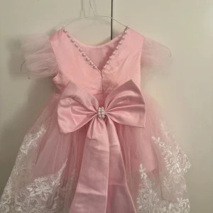 Rosa prinsessklänning med pärlor och rosett - Superfin rosa klänning med puffiga tyllärmar, pärlor längs v-ringningen och stor rosett med pärldetalj framtill. Kjolen är dekorerad med vit broderad spets och har en fluffig, drömmig look. Perfekt för dig som vill sticka ut med en söt och elegant stil.