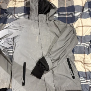 Grå vindjacka med huva  - Säljer en grå vindjacka med huva och svarta detaljer vid ärmslut. Jackan har två fickor med dragkedja framtill och är perfekt för blåsiga dagar. Materialet är lätt och syntetiskt, vilket gör den smidig att bära. Enkel och stilren design som funkar till många outfits.