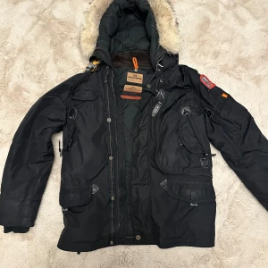 Svart Parajumpers parkajacka med päls - Svart parkajacka från Parajumpers med huva och beige fuskpäls, flera fickor med dragkedja och knappar, samt ikonisk gul rem vid kragen. Jackan har patch på ärmen och detaljer i orange. Perfekt för kalla dagar med snygg streetstil.