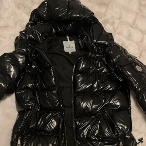 Svart pufferjacka från Moncler - Säljer en svart, glansig pufferjacka från Moncler med huva och dragkedja framtill. Jackan har quiltad design, fickor med dragkedja och en Moncler-logga på ärmen. Perfekt för kalla dagar och har en riktigt snygg streetstil. Ombyte