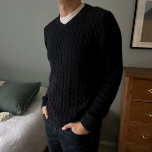 Mörkblå kabelstickad tröja V-ringad - Snygg mörkblå kabelstickad tröja från Bondelid med V-ringning och långa ärmar. Tröjan har ett klassiskt mönster med grova flätor och ribbade muddar vid ärmslut och nederkant. Perfekt för dig som gillar stilrena och tidlösa plagg.