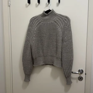 Ljusgrå stickad polotröja från H&M - Säljer en chunky grå stickad polotröja från H&M med grov ribbad struktur och hög krage. Tröjan har långärmade ballongärmar och breda muddar som ger en cool och avslappnad vibe. Perfekt för att hålla sig varm under kyliga dagar.