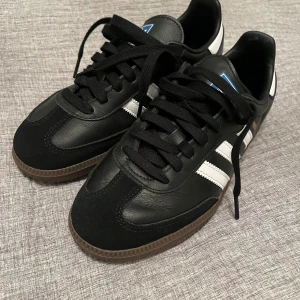 Adidas Samba svart/vit strl 39 1/3 - Klassiska Adidas Samba sneakers i svart och vitt med bruna gummisulor. Skorna har de ikoniska tre vita ränderna på sidan, svart ovandel i skinn och mocka samt vit hälkappa. 