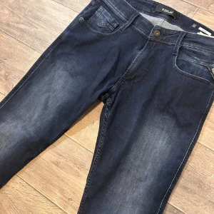Replay Anbass dark indigo jeans - Snygga mörkblå Replay Anbass jeans med klassisk femficksdesign och diskreta slitningar. Jeansen har raka ben och normal passform, tillverkade i mjukt denimtyg med stretch för extra komfort. Perfekta för en avslappnad och stilren look.