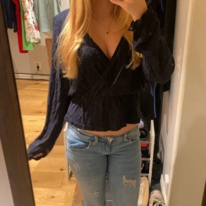 Marinblå omlott blus med volang - Säljer en marinblå omlott blus med lång ärm och v-ringad hals. Blusen har en snygg volangdetalj vid midjan och är croppad i modellen. Materialet är lätt och har en lite skrynklig struktur som ger en cool look. Perfekt att styla med jeans eller kjol. Storlek L men skulle säga att den passar s-m mer då jag är storlek xs och sitter fint på mig