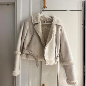 Beige teddyjacka från Miss Selfridge - Snygg beige teddyjacka från Miss Selfridge i storlek 38. Jackan har mjukt foder, bred krage, dragkedja och bälte i midjan. Fuskpälsdetaljer vid ärmslut och nederkant ger en extra mysig känsla. Perfekt för kyliga dagar!