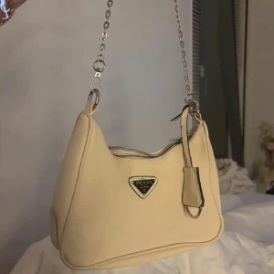 Beige Axelväska - Snygg beige axelväska med klassisk silverfärgad kedja och dragkedja upptill. Väskan har en stilren, mjuk form och ikonisk logga framtill. Insidan är fodrad med svart tyg med diskret mönster och metallbricka med märkning.🫶🏻