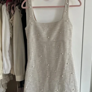Beige pärlprydd kortklänning - Supersöt beige kortklänning med pärldetaljer över hela klänningen. Klänningen har breda axelband och en rak, något figurnära passform. Materialet är strukturerat och ger en lyxig känsla. Perfekt för dig som gillar stilrena och eleganta plagg med det lilla extra.