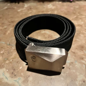 Svart tygbälte Sail Racing - Snyggt svart bälte i slitstarkt tyg med ett coolt, geometriskt silverfärgat metallspänne. Spännet har en unik design med en liten logotyp ingraverad. . Den är 117 cm lång inklusive allt. Ny pris för bältet är 499 kr 