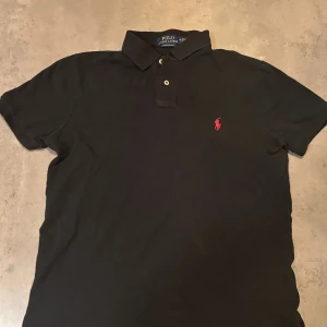 Svart piké från Polo Ralph Lauren - Svart pikétröja från Polo Ralph Lauren i slim fit med klassisk krage och två knappar framtill. Den har det ikoniska röda broderade Polo-logot på bröstet. Materialet är mjuk bomull som känns skön mot huden och passar perfekt till jeans eller shorts. Bara använd två gånger 