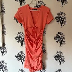 Orange kort klänning från SHEIN - Snygg Orange kort klänning från SHEIN med djup v-ringning, snörning framtill och rynkade detaljer längs kjolen. Klänningen har korta ärmar och är figurnära med volangkant nertill. Perfekt för dig som vill sticka ut med en färgstark look.