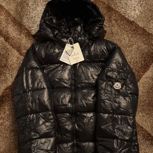 Svart Moncler pufferjacka Xl - Svart pufferjacka från Moncler i storlek XL med huva och klassisk Moncler-logga på ärmen. Jackan har dragkedja framtill och quiltad design. Insidan har en cool serietidningsetikett. Perfekt för dig som vill ha en snygg och varm jacka med streetkänsla.mycket vacker 