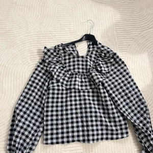 Svartvit rutig blus med volang - Säljer en svartvit rutig blus med långärmade puffärmar och bred volang runt halsringningen. Blusen har en klassisk rutig design och är tillverkad i ett lätt bomullstyg som känns skönt mot huden. Perfekt för dig som gillar statement-detaljer och trendiga mönster.